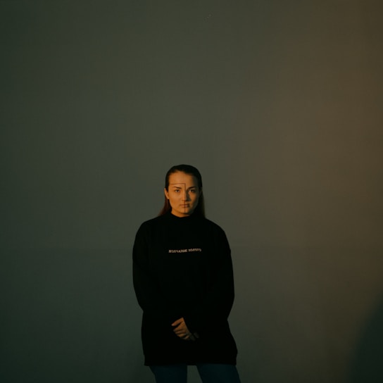 A person stands in a dark room with minimal lighting, wearing a black sweatshirt with white text. The background is a plain, dark green wall creating a mysterious and somber atmosphere. The person has minimal facial expressions and appears to be posing calmly with arms crossed at the waist.