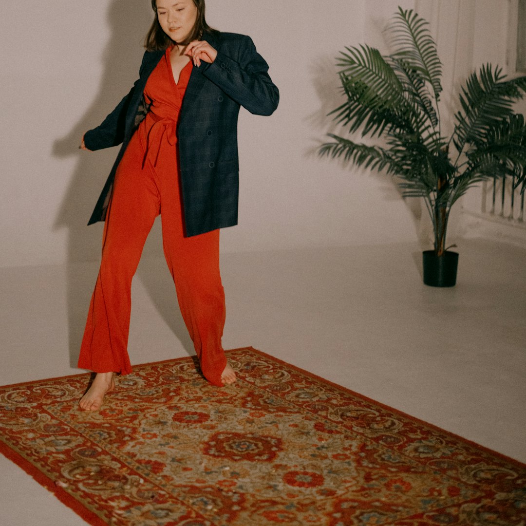 woman in black coat and orange pants standing on red and brown floral area rug