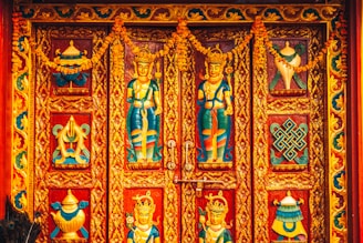 Intricately designed wooden doors featuring vibrant carvings of deities and ornate patterns, bordered by decorative floral motifs and adorned with garlands.