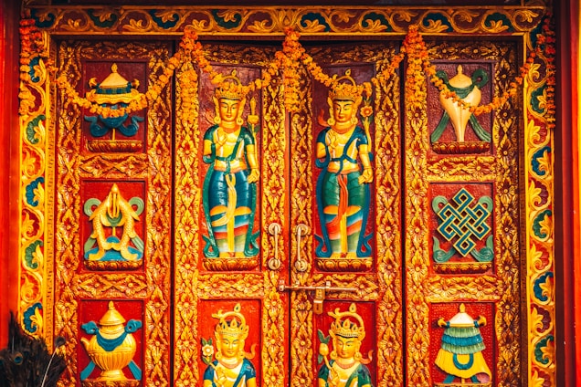 Intricately designed wooden doors featuring vibrant carvings of deities and ornate patterns, bordered by decorative floral motifs and adorned with garlands.