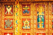 Intricately carved and vibrantly painted wooden panel featuring religious and cultural motifs, including a deity figure and geometric patterns surrounded by floral designs. The dominant colors are rich reds, blues, yellows, and greens, contributing to a lively and ornate aesthetic.