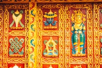 Intricately carved and vibrantly painted wooden panel featuring religious and cultural motifs, including a deity figure and geometric patterns surrounded by floral designs. The dominant colors are rich reds, blues, yellows, and greens, contributing to a lively and ornate aesthetic.