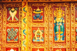Intricately carved and vibrantly painted wooden panel featuring religious and cultural motifs, including a deity figure and geometric patterns surrounded by floral designs. The dominant colors are rich reds, blues, yellows, and greens, contributing to a lively and ornate aesthetic.
