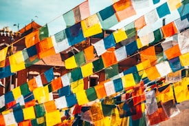 Brightly colored prayer flags are strung across an outdoor setting, fluttering in the wind. The flags are primarily blue, white, red, green, and yellow, with intricate symbols and text printed on them. The background features parts of rooftops and elements typical of an urban landscape under a clear blue sky.