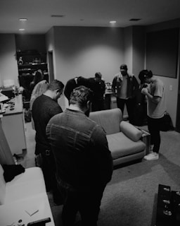 A group of individuals bow their heads in a small room, possibly engaged in a moment of silence or prayer. The scene appears casual with a sofa and various items scattered around, suggesting a private or backstage area.