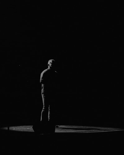 A person stands alone on a dimly lit stage, surrounded by darkness. The lighting creates a dramatic and contemplative atmosphere, highlighting the individual's silhouette.