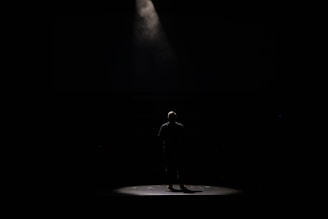 A lone figure stands under a spotlight on a dark stage, creating a dramatic contrast between the light and the surrounding darkness.