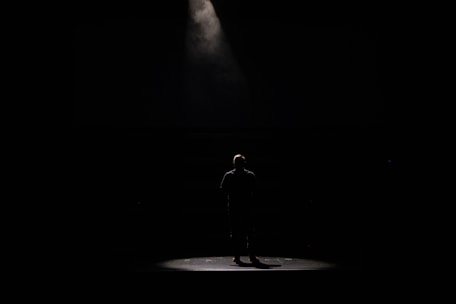 A lone figure stands under a spotlight on a dark stage, creating a dramatic contrast between the light and the surrounding darkness.