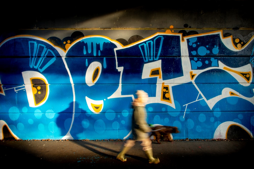 Femme en chemise grise et pantalon noir debout à côté d’un mur bleu et jaune