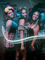 Three women dressed in costumes stand side by side, smiling at the camera. The woman on the left has face paint resembling a sugar skull and flowers in her hair. The middle woman wears cat ears and a leopard print outfit. The woman on the right sports a black dress with a corset-like design. A variety of colorful lights and balloons are visible in the background.