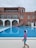 A person dressed in a swimming outfit stands by the side of an outdoor swimming pool. The person appears to be a child wearing a pink and blue swimsuit with a swim cap. In the background, there is a large building with multiple windows and a circular staircase leading down to the pool area. There are also pool benches and umbrellas near the building, and the sky is overcast.