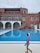 A person dressed in a swimming outfit stands by the side of an outdoor swimming pool. The person appears to be a child wearing a pink and blue swimsuit with a swim cap. In the background, there is a large building with multiple windows and a circular staircase leading down to the pool area. There are also pool benches and umbrellas near the building, and the sky is overcast.