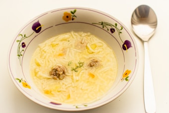A bowl of creamy soup with small pasta and visible meat pieces, garnished with a sprig of dill. The bowl has a floral design with purple and yellow flowers, accompanied by a metal spoon set on a white surface.