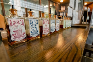A row of square glass bottles of FEW whiskey is displayed on a wooden counter. The bottles have distinct labels featuring different designs and colors, including red and blue, with text detailing the type of whiskey. The setting appears to be a bar or distillery, with a warm, inviting atmosphere enhanced by soft lighting.