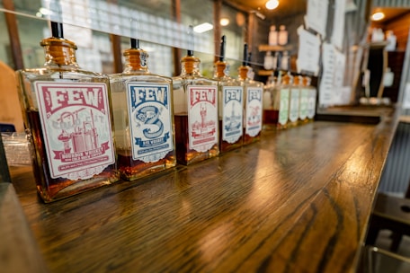 A row of square glass bottles of FEW whiskey is displayed on a wooden counter. The bottles have distinct labels featuring different designs and colors, including red and blue, with text detailing the type of whiskey. The setting appears to be a bar or distillery, with a warm, inviting atmosphere enhanced by soft lighting.