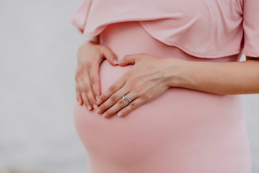 Gestante acariciando a barriga com as mãos em formato de coração, vestido rosa