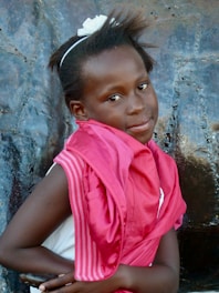 A young girl with short hair and a white headband is standing against a textured, metallic surface. She is wearing a pink jacket draped over her shoulders and a white top. Her gaze is directed towards the camera with a slight smile.