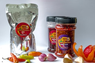 Three containers of sambal, a type of Indonesian chili sauce, are prominently displayed. One is a metallic pouch with a label, and the other two are transparent jars with black lids, both labeled. Around them are fresh ingredients like chili peppers, onions, and garlic, scattered on a white surface.