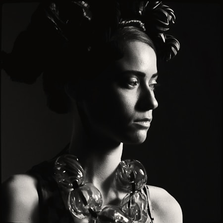 A monochrome portrait of a woman looking to the right, showcasing strong chiaroscuro lighting. Her hair is styled elaborately with large adornments. She wears a necklace of large, rounded beads, creating a contrast with her skin tone. The image highlights textures and shadows on her face and the decorative items.