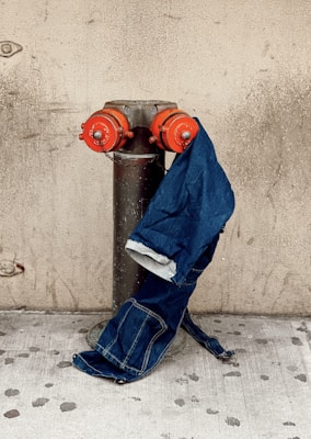 A pair of blue jeans is draped over a fire hydrant with red caps, set against a textured beige wall. The concrete pavement below the hydrant is slightly stained, complementing the industrial urban aesthetic.