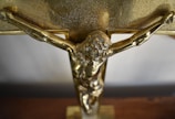 A close-up view of a golden crucifix with a detailed depiction of Jesus Christ on the cross. The surface of the cross appears textured with a shiny metallic finish.