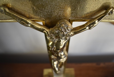 A close-up view of a golden crucifix with a detailed depiction of Jesus Christ on the cross. The surface of the cross appears textured with a shiny metallic finish.