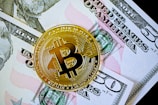 A gold-colored Bitcoin coin is placed on top of a fifty-dollar bill. The Bitcoin features the letter 'B' with intricate circuit-like patterns along its surface, symbolizing digital currency.