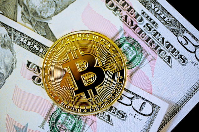 A gold-colored Bitcoin coin is placed on top of a fifty-dollar bill. The Bitcoin features the letter 'B' with intricate circuit-like patterns along its surface, symbolizing digital currency.