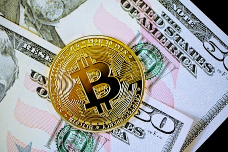 A gold-colored Bitcoin coin is placed on top of a fifty-dollar bill. The Bitcoin features the letter 'B' with intricate circuit-like patterns along its surface, symbolizing digital currency.