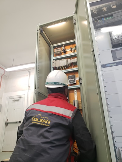 A person wearing a hard hat and a red and gray jacket with reflective stripes is working on an open electrical cabinet filled with various components and wires. The environment appears to be industrial with fluorescent lighting.