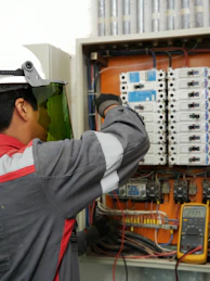 An electrician in a hard hat and face shield testing an electrical panel