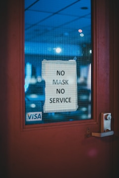 A sign taped to a glass door reads 'NO MASK NO SERVICE'. The background is slightly blurred, showing indoor lights and a reflection. A Visa sticker is seen in the lower left corner of the sign, and the door appears to be red with a modern silver door handle.