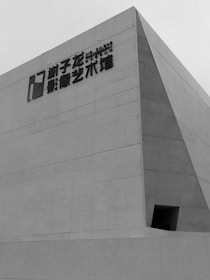 A modern concrete structure with angular lines and a minimalist design. The building features an exterior with large, smooth surface panels interrupted by uniform dots. Chinese characters and a logo are mounted on the wall near the top, indicating it's an artistic or cultural institution.