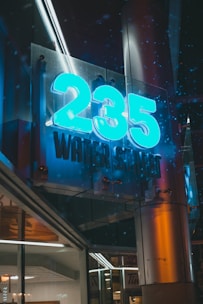 A glowing sign displaying '235 WATER STREET' with bright blue numbers and text, mounted on a building facade. The scene includes reflections in the glass and metal surfaces, contributing to a modern urban aesthetic. It appears to be night time, with a dimly lit street visible in the background.