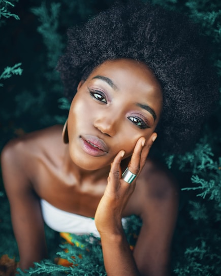 A person with natural curly hair is gazing directly at the camera, with one hand resting gently on their cheek. The background is filled with lush green foliage, providing a contrast to the person's dark hair and smooth skin. They are wearing a strapless top and a silver ring, with a calm and serene expression.