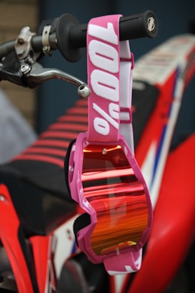 A pair of pink goggles with reflective lenses hangs from the handlebar of a motorcycle. The strap of the goggles features the text '100%' in white. The motorcycle has a red and black color scheme with a visible grip and brake lever.