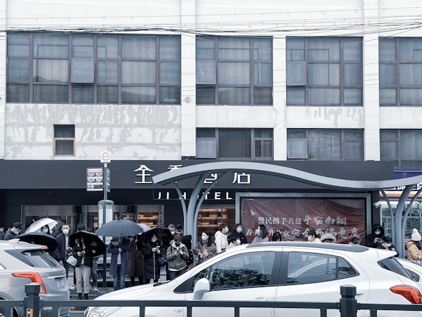 People are standing in line under umbrellas in front of a building. Cars are parked in the foreground. The building has a large sign with text on it and many windows.