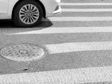 A car tire is partially visible on a striped pedestrian crosswalk beside a round manhole cover on an asphalt surface.