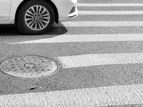 A car tire is partially visible on a striped pedestrian crosswalk beside a round manhole cover on an asphalt surface.