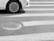 A car tire is partially visible on a striped pedestrian crosswalk beside a round manhole cover on an asphalt surface.