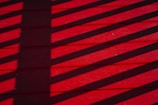 Shadows of a fence or railing cast diagonal stripes on a vivid red wooden deck. The shadows create a contrasting pattern across the surface, adding depth to the horizontal planks.