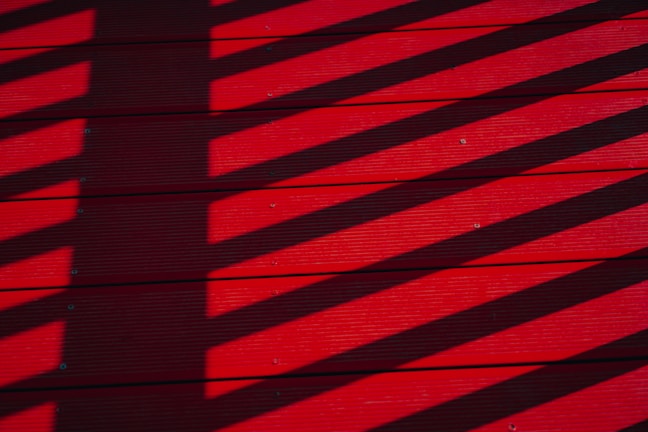 Shadows of a fence or railing cast diagonal stripes on a vivid red wooden deck. The shadows create a contrasting pattern across the surface, adding depth to the horizontal planks.