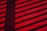 Shadows of a fence or railing cast diagonal stripes on a vivid red wooden deck. The shadows create a contrasting pattern across the surface, adding depth to the horizontal planks.