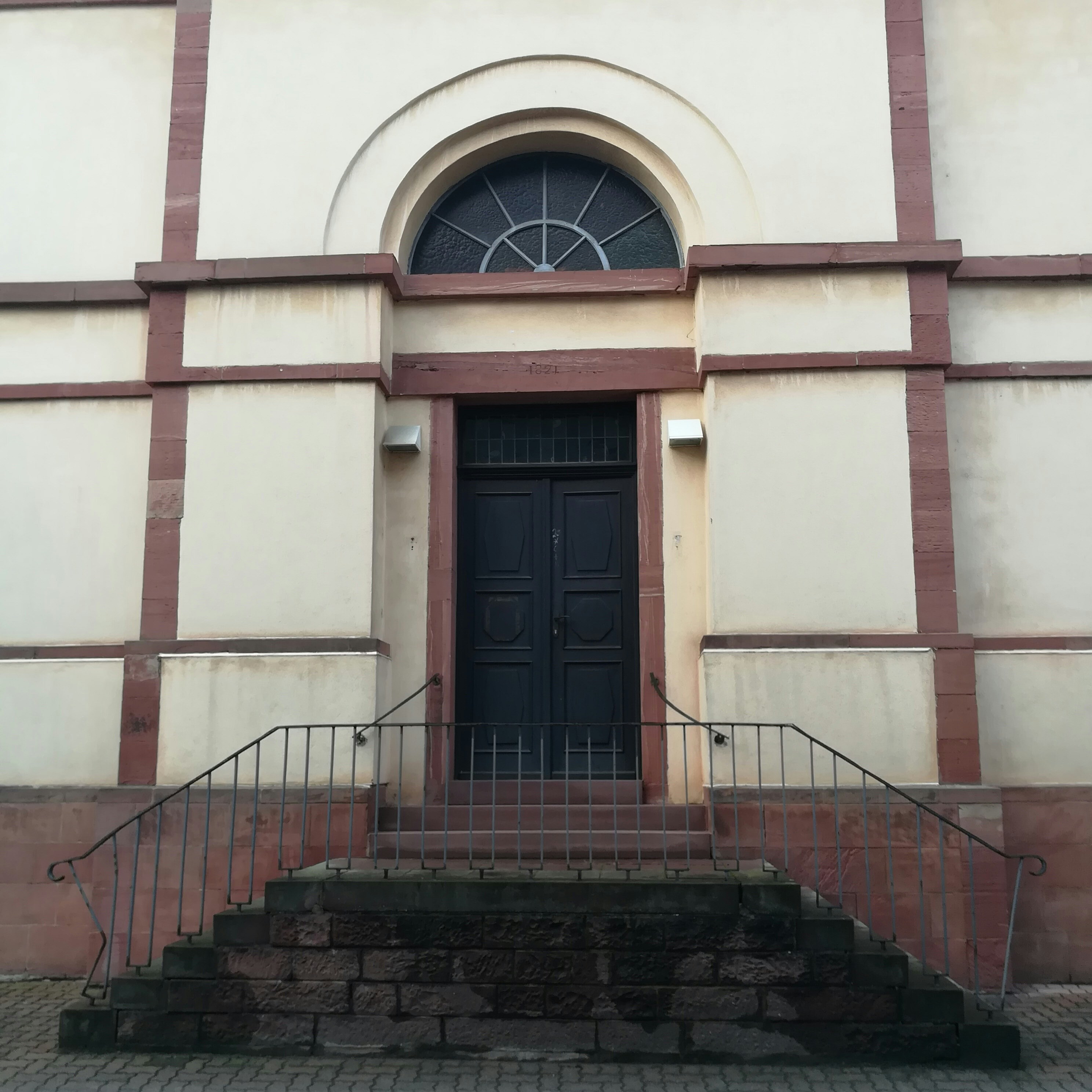 A dark wooden door is set within a beige wall framed by red brick accents. The door is accessed by a short set of stone steps with metal railings on either side. Above the door is an arched window with a decorative fanlight design. The building exterior appears to be historic and solidly constructed.