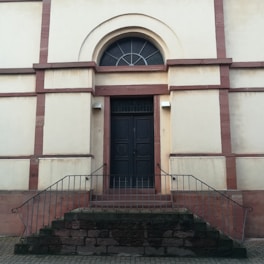 A dark wooden door is set within a beige wall framed by red brick accents. The door is accessed by a short set of stone steps with metal railings on either side. Above the door is an arched window with a decorative fanlight design. The building exterior appears to be historic and solidly constructed.