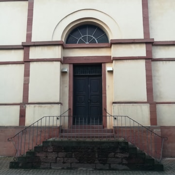 A dark wooden door is set within a beige wall framed by red brick accents. The door is accessed by a short set of stone steps with metal railings on either side. Above the door is an arched window with a decorative fanlight design. The building exterior appears to be historic and solidly constructed.