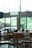 A spacious cafe interior with wooden tables and chairs. Large windows let in natural light, creating a bright and airy atmosphere. Several plants are placed on the tables, enhancing the cozy ambiance. Outside, greenery is visible, and there is a parked car.