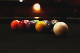 A row of billiard balls aligned on a dark pool table, with low ambient lighting creating a warm, cozy atmosphere. The balls are in various colors, including red, purple, yellow, and white, and are positioned diagonally towards the viewer. In the background, a blurred light source adds to the dim, intimate setting.