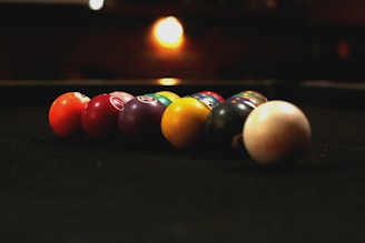 A row of billiard balls aligned on a dark pool table, with low ambient lighting creating a warm, cozy atmosphere. The balls are in various colors, including red, purple, yellow, and white, and are positioned diagonally towards the viewer. In the background, a blurred light source adds to the dim, intimate setting.