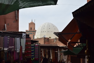 A lively marketplace scene with colorful fabrics hanging from stalls in the foreground. A large, white dome and a minaret are visible in the background, surrounded by earthy-toned buildings. The stalls are partially covered by makeshift roofing, creating a cozy and bustling atmosphere.